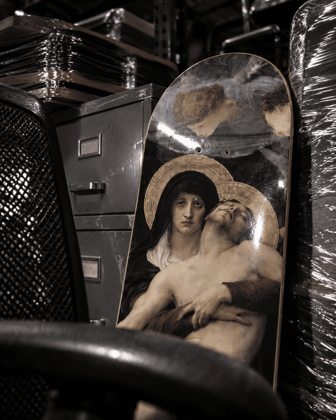 Bouguereau Pieta skateboard deck leaning against a filing cabinet in a warehouse storage room