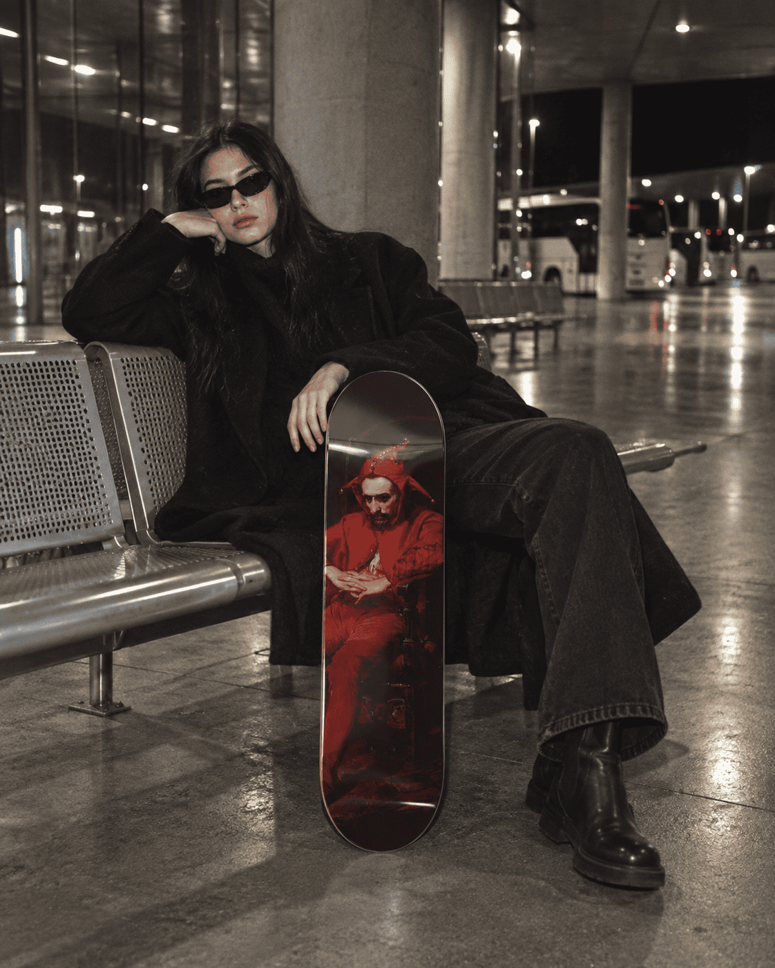 Person in black holding a skateboard deck with a red-robed melancholy jester portrait at a night bus station