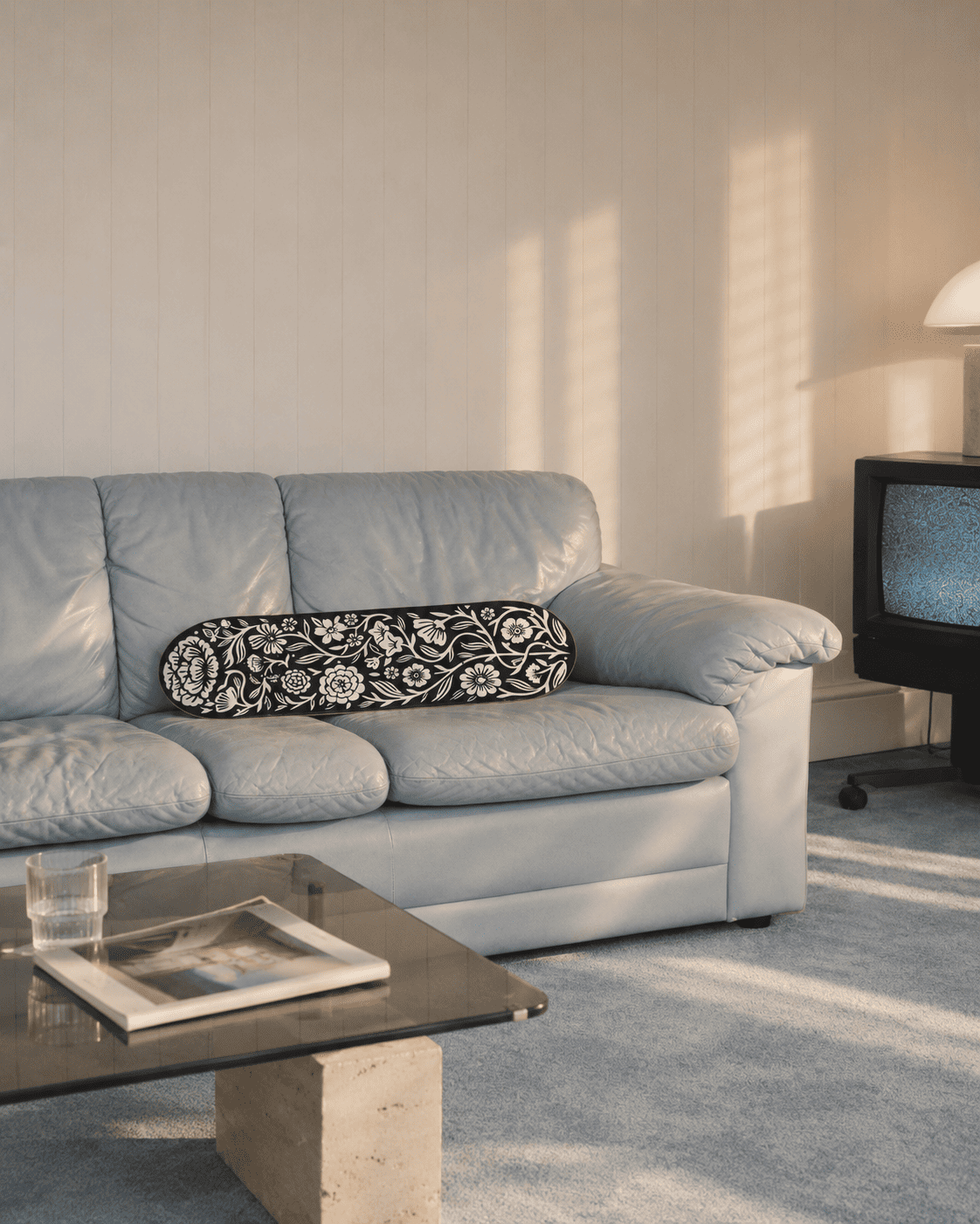 Black and white floral wall art on a skateboard deck lying across the cushions of a pale blue-grey leather sofa in warm late-afternoon light, a glass-and-travertine coffee table with a magazine in front and a CRT television at the side