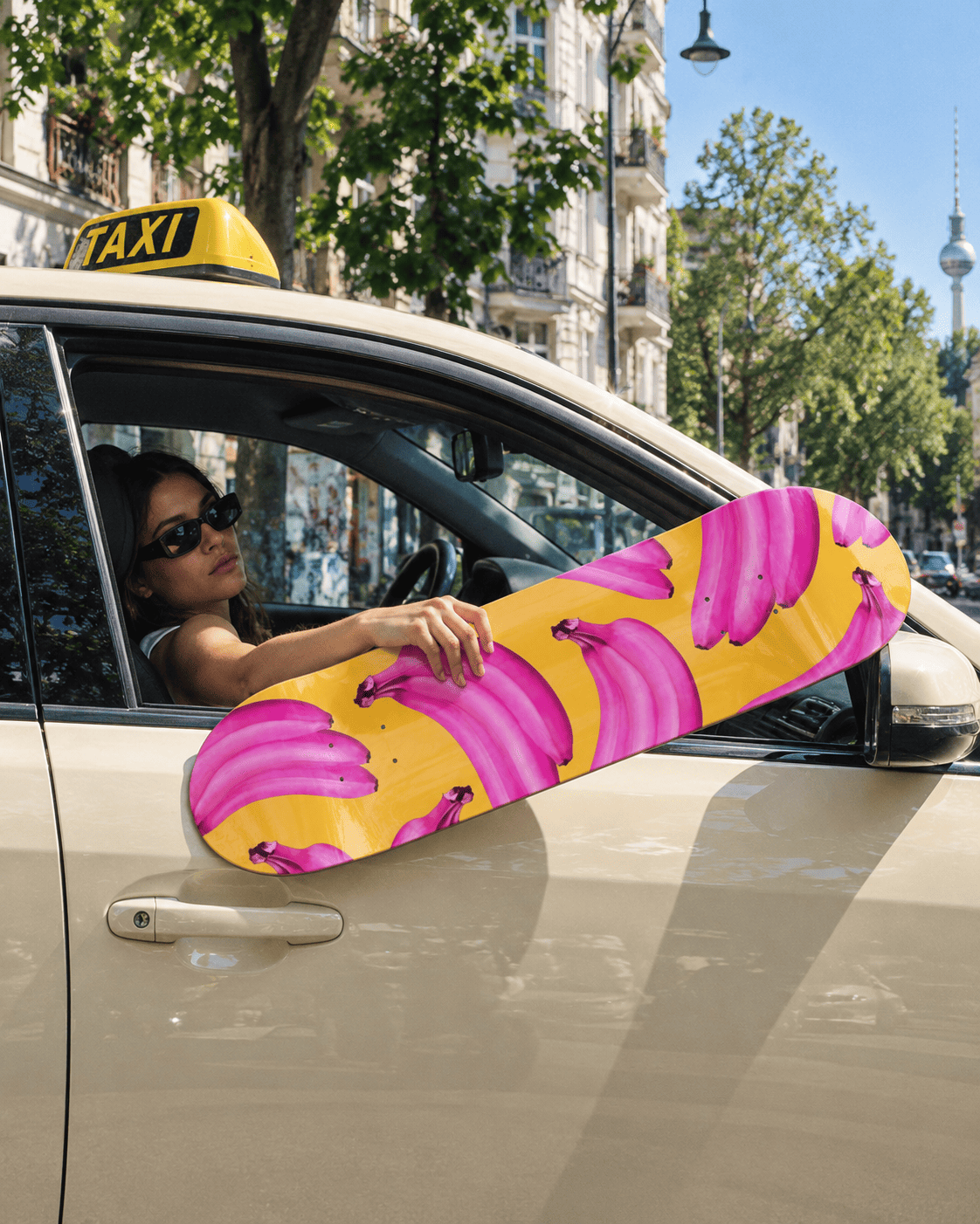 A take on the banana in pop art: a skateboard deck with hot-pink bananas on yellow held out of the driver's window of a cream taxi with a yellow TAXI roof sign on a sunny tree-lined street